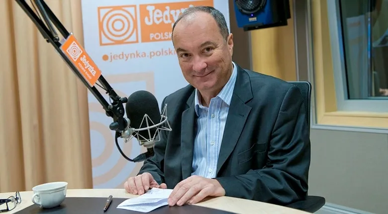 Mężczyzna w marynarce uśmiecha się, siedząc w studio radiowym przy mikrofonie, z notatkami i filiżanką na stole oraz widocznym logo stacji w tle.
