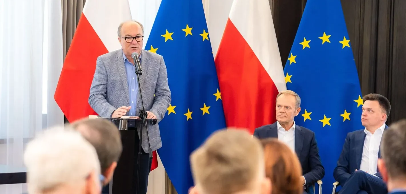 Mężczyzna przemawia z mównicy na tle flag Polski i Unii Europejskiej, podczas gdy dwóch mężczyzn w garniturach siedzi obok.