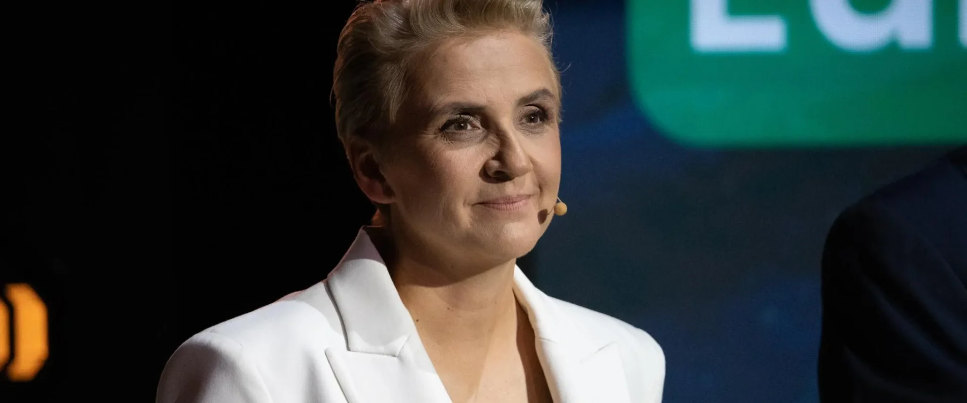 Kobieta o krótkich blond włosach w białym żakiecie stoi na scenie z mikrofonem na ucho, uśmiechnięta, na tle ciemnego planu i zielonego ekranu z fragmentem napisu EU.