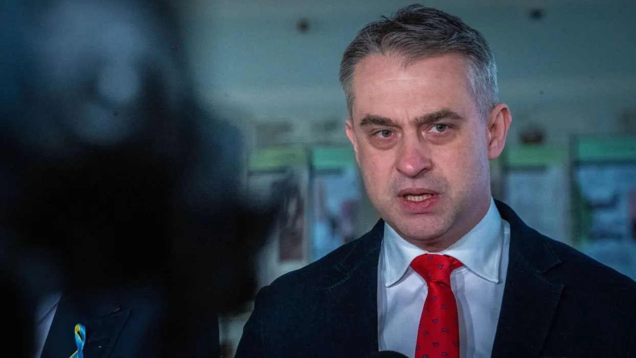 Mężczyzna w garniturze z czerwonym krawatem przemawia do mediów, wyrażając emocje w poważnej atmosferze.