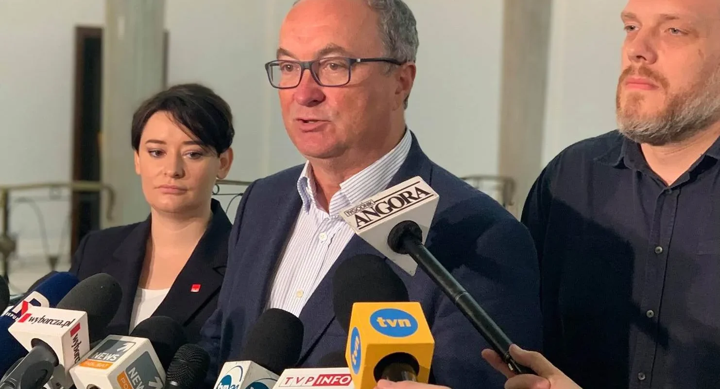 Mężczyzna w garniturze i okularach przemawia do mediów podczas konferencji prasowej, otoczony mikrofonami i dwoma współpracownikami.