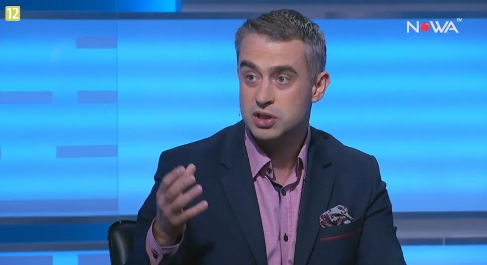 Mężczyzna w marynarce i koszuli energicznie gestykuluje i mówi w studiu telewizyjnym na niebieskim tle, z widocznym logo stacji w prawym górnym rogu.