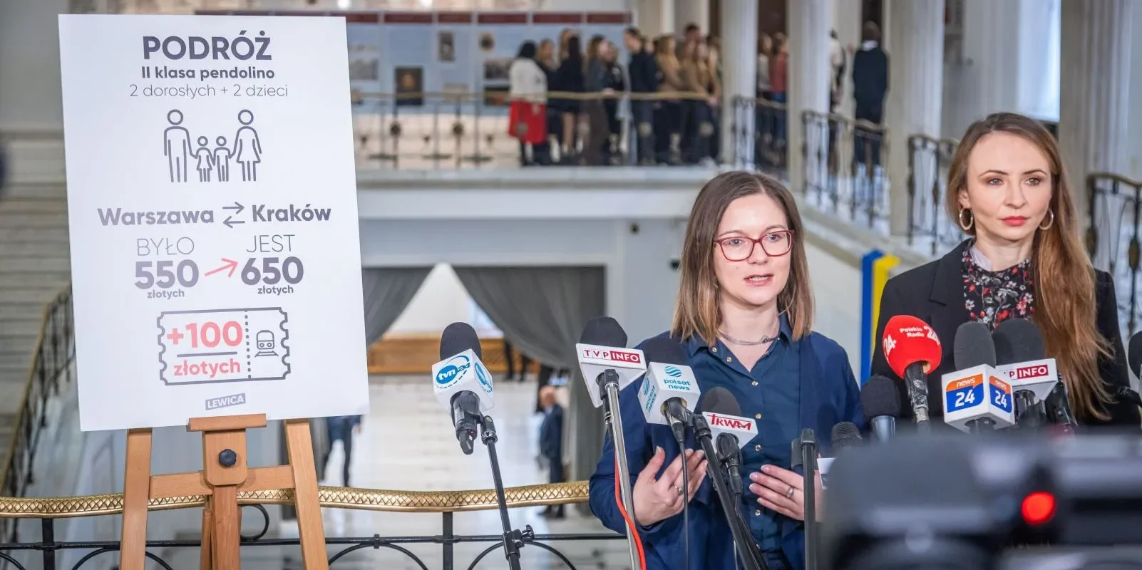 Na zdjęciu widoczne są dwie kobiety podczas konferencji prasowej dotyczącej zmian w cenach biletów kolejowych między Warszawą a Krakowem, z plakatem informacyjnym w tle.