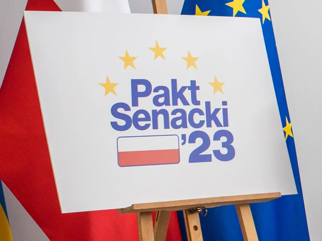 Plakat na sztaludze z napisem „Pakt Senacki ’23”, otoczony żółtymi gwiazdami, stoi między flagą Polski a flagą Unii Europejskiej.