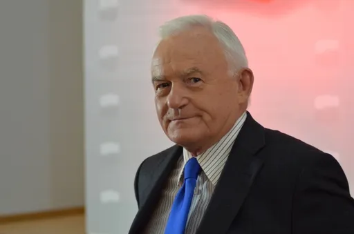 Starszy mężczyzna w czarnym garniturze i niebieskim krawacie siedzi w jasnym wnętrzu i patrzy w kamerę.