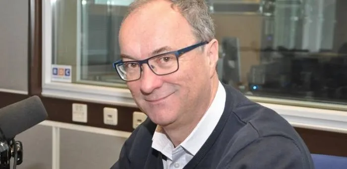 Mężczyzna w okularach siedzi przy mikrofonie w radiowym studiu i lekko się uśmiecha.