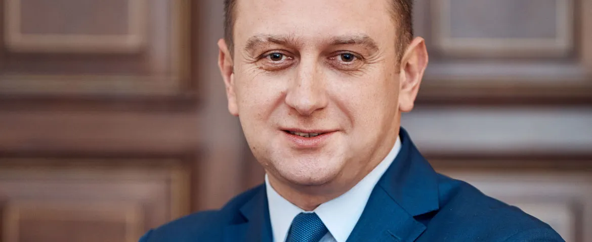 Półzbliżeniowy portret uśmiechniętego mężczyzny w granatowym garniturze i niebieskim krawacie na tle zdobionych drewnianych drzwi.