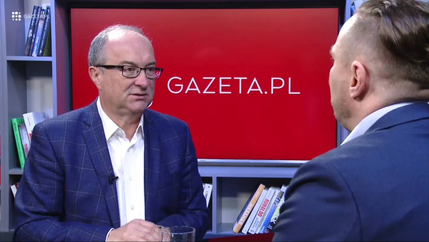 Dwóch mężczyzn rozmawia w studiu telewizyjnym; po lewej siedzi w okularach w granatowym żakiecie, na tle czerwnego ekranu Gazeta.pl i półek z książkami.