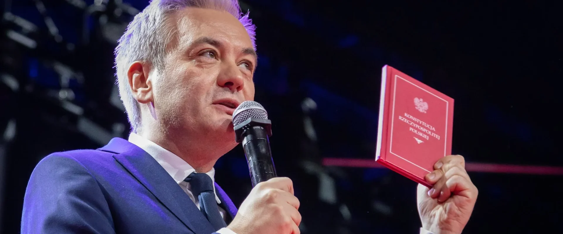Mężczyzna w granatowym garniturze przemawia na scenie, trzymając w jednej ręce mikrofon, a w drugiej czerwony podręcznik Konstytucji RP.