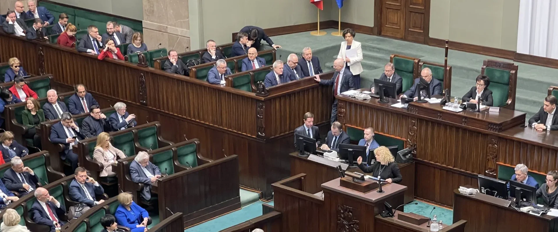 Na zdjęciu widać posiedzenie parlamentu, z przedstawicielami różnych ugrupowań politycznych siedzącymi w drewnianych ławach.