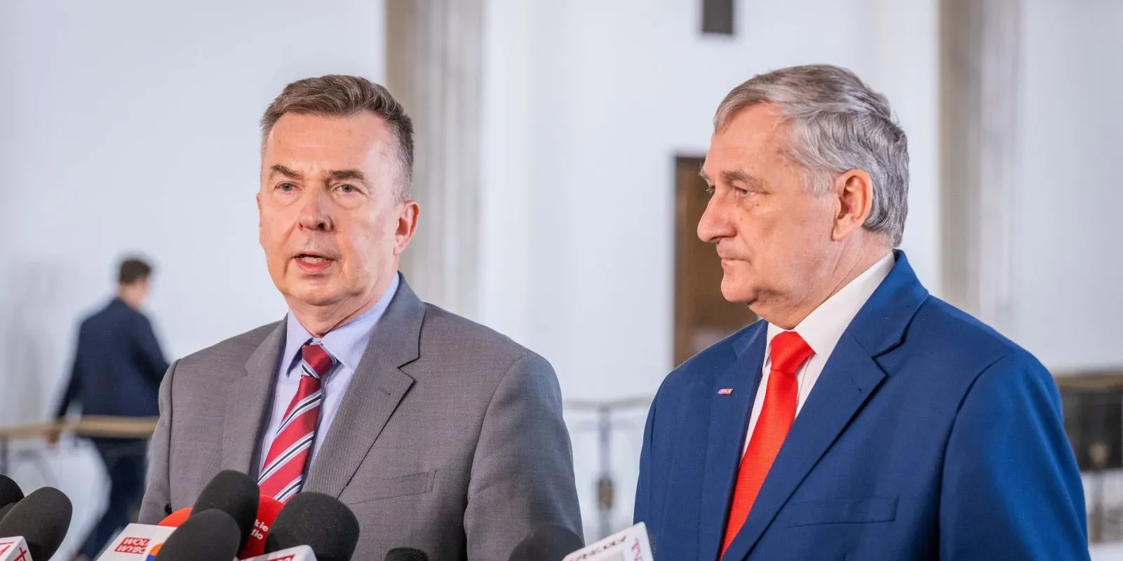 Dwóch mężczyzn w garniturach stoi podczas konferencji prasowej z licznymi mikrofonami, omawiając wyższe świadczenia dla emerytów mundurowych, którzy pracowali i płacili ZUS w cywilu.
