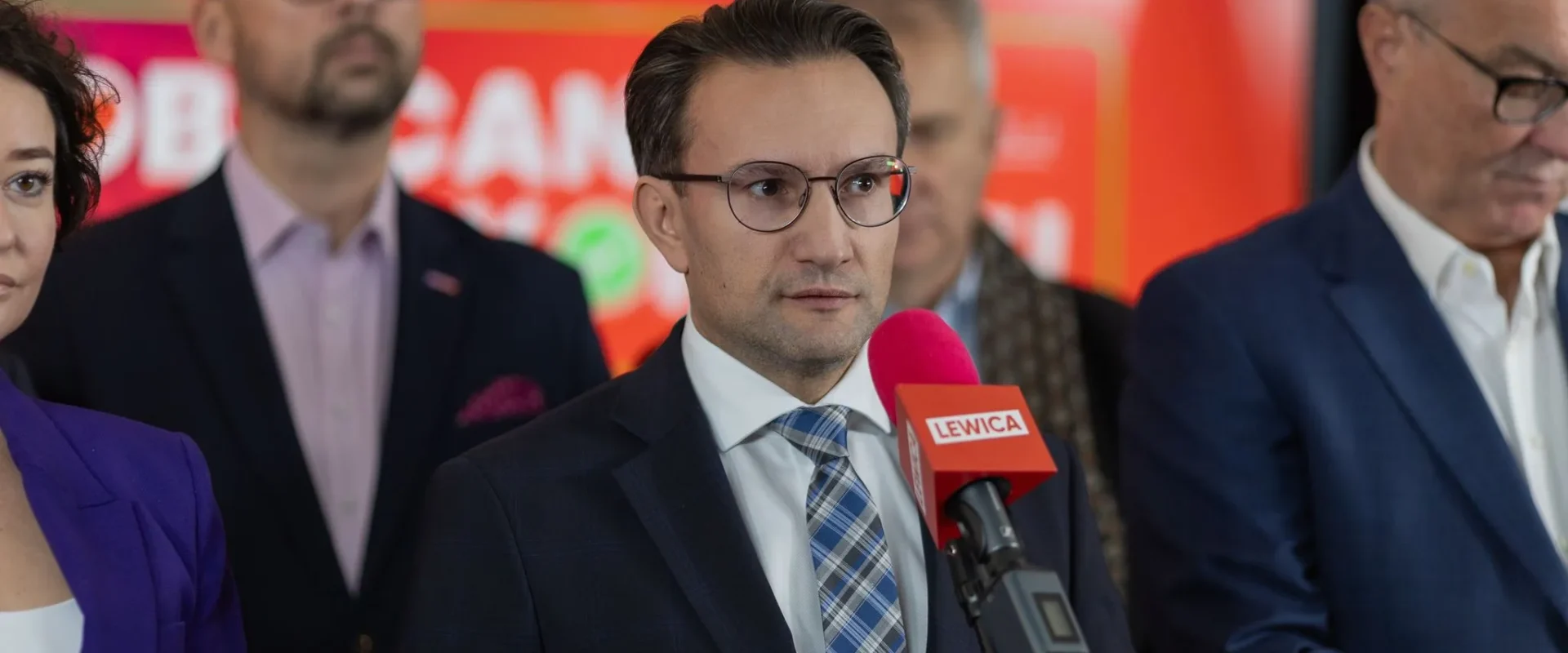 Mężczyzna w garniturze i okularach przemawia do mikrofonu z logiem Lewica podczas konferencji prasowej, w tle stoją inni politycy na czerwono-pomarańczowym tle.