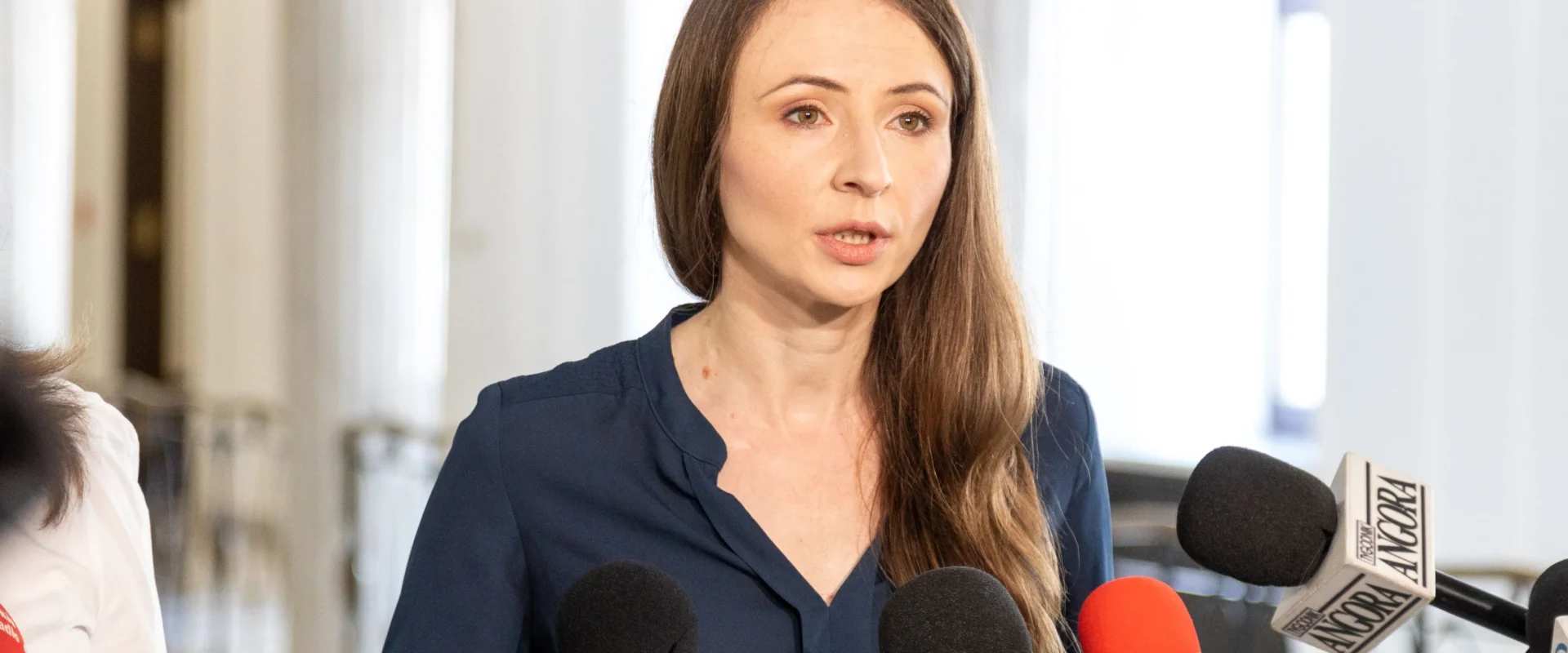Posłanka Lewicy przemawia do mediów podczas konferencji prasowej, stojąc przy mikrofonach.