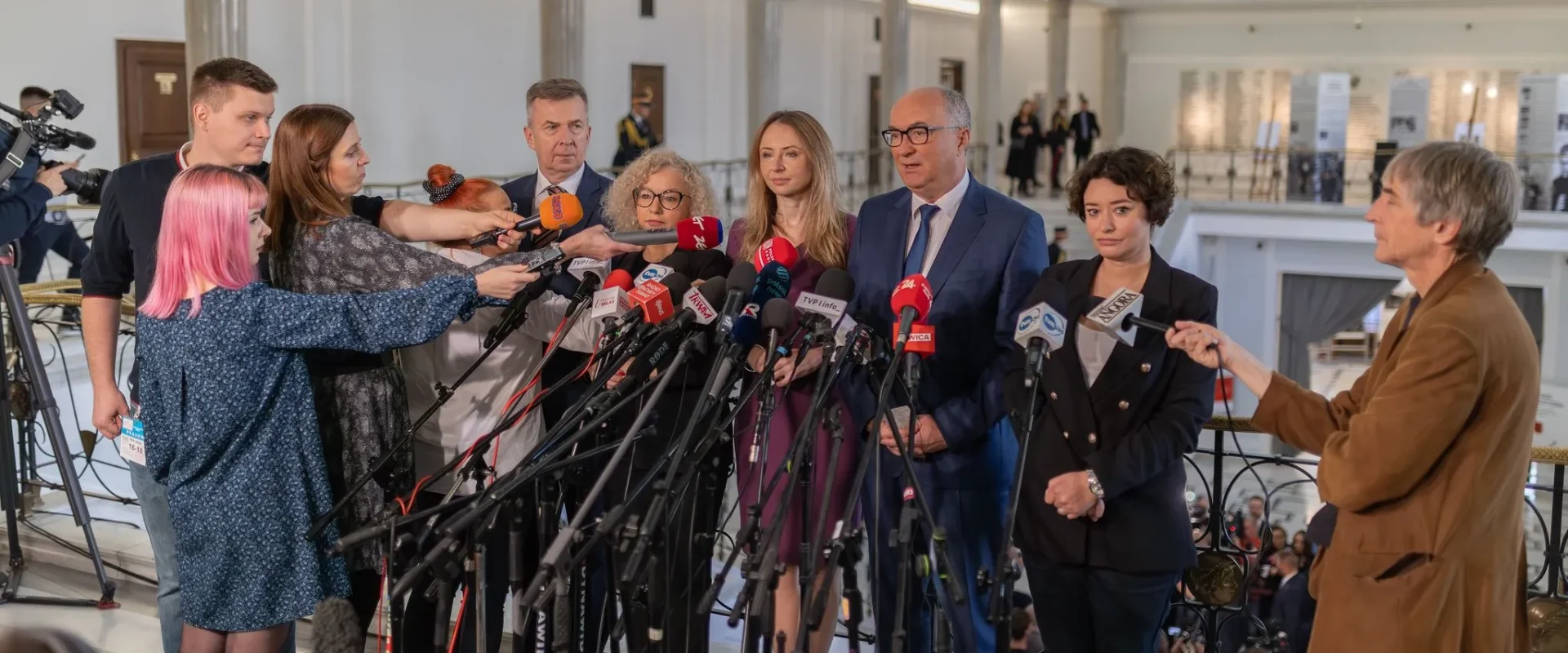 Grupa urzędników w garniturach udziela konferencji prasowej, otoczona licznymi mikrofonami i reporterami.