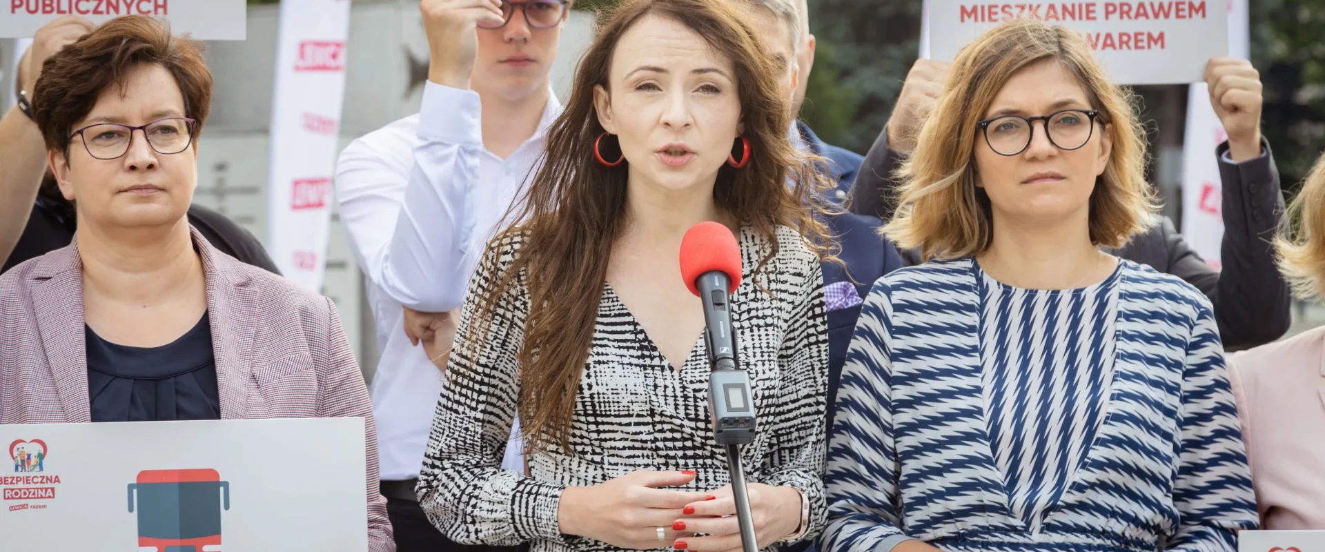 Na zdjęciu widać grupę osób podczas konferencji prasowej, gdzie jedna z kobiet przemawia do mikrofonu, a pozostali trzymają plakaty z hasłami dotyczącymi polityki społecznej.