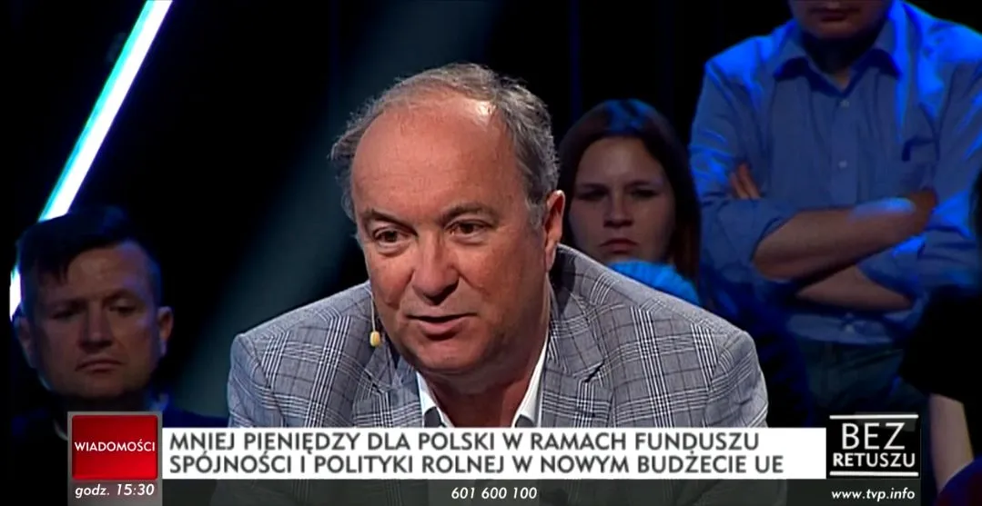 Mężczyzna w garniturze z mikrofonem na uchu przemawia w studiu telewizyjnym, z napisem informacyjnym na dole ekranu.