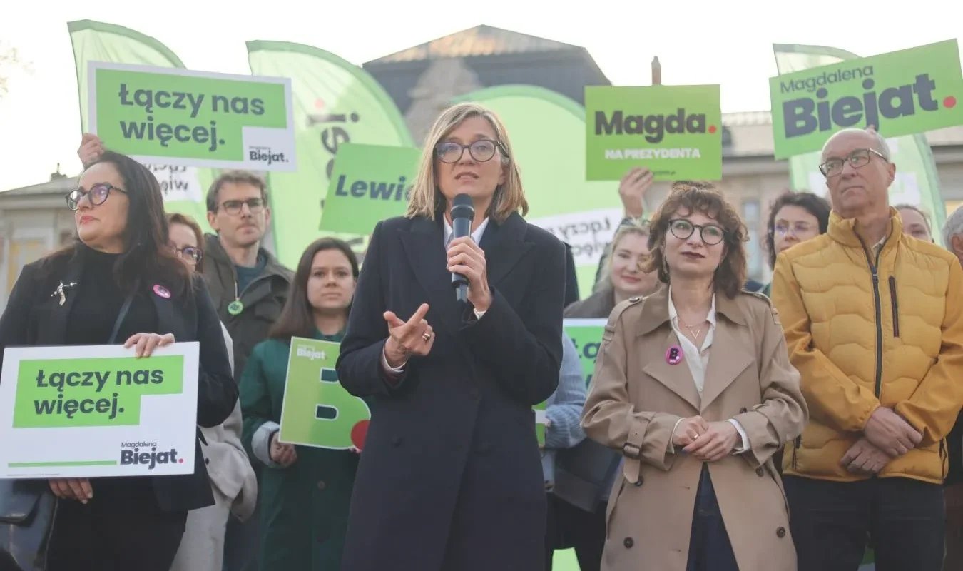 Grupa osób na wiecu wyborczym, przed mikrofonem przemawia kobieta w czarnym płaszczu, w otoczeniu zielonych banerów i plakatów z hasłem „Łączy nas więcej”.