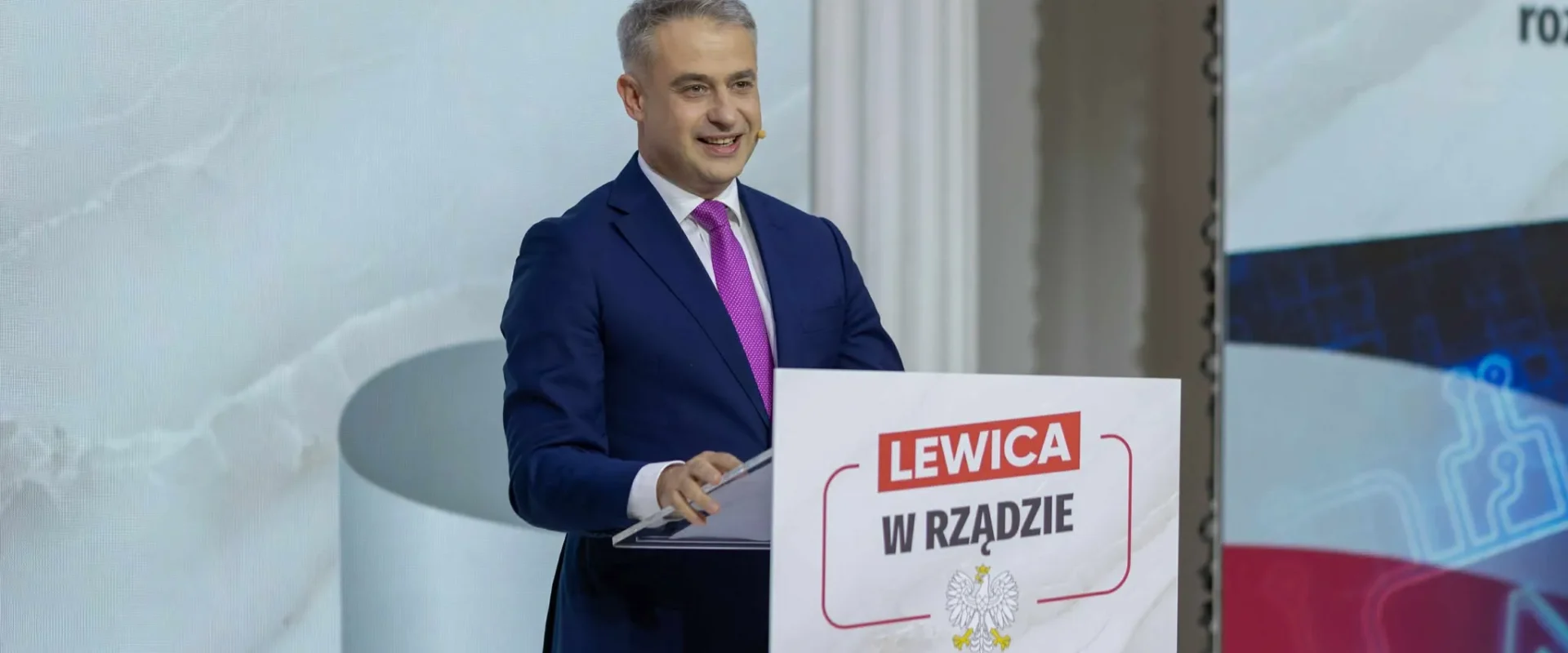 Mężczyzna w granatowym garniturze i różowym krawacie przemawia ze sceny przy mównicy z napisem „LEWICA W RZĄDZIE”.