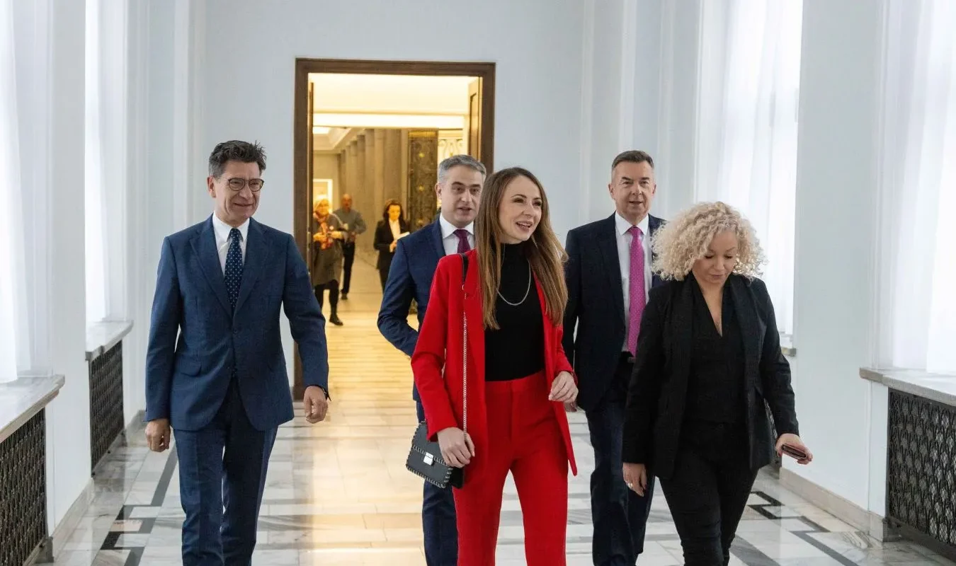 Grupa osób w garniturach spaceruje po eleganckim, jasnym korytarzu; na pierwszym planie kobieta ubrana w czerwony garnitur prowadzi grupę.