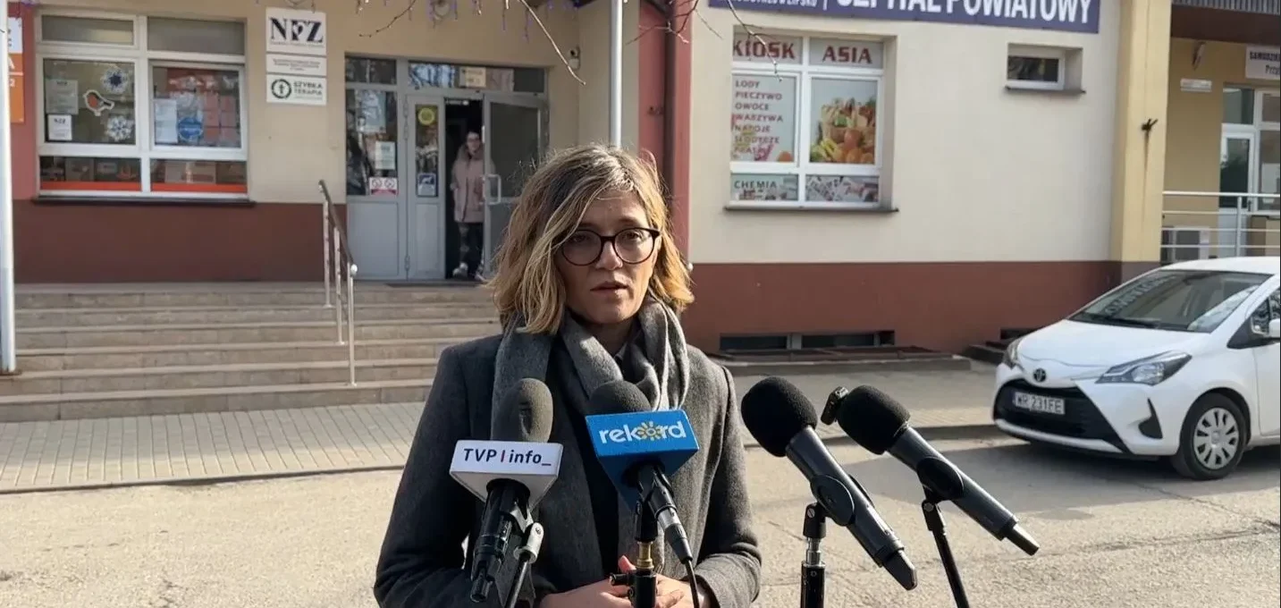 Kobieta z okularami przemawia do mediów podczas konferencji prasowej przed wejściem do szpitala powiatowego.