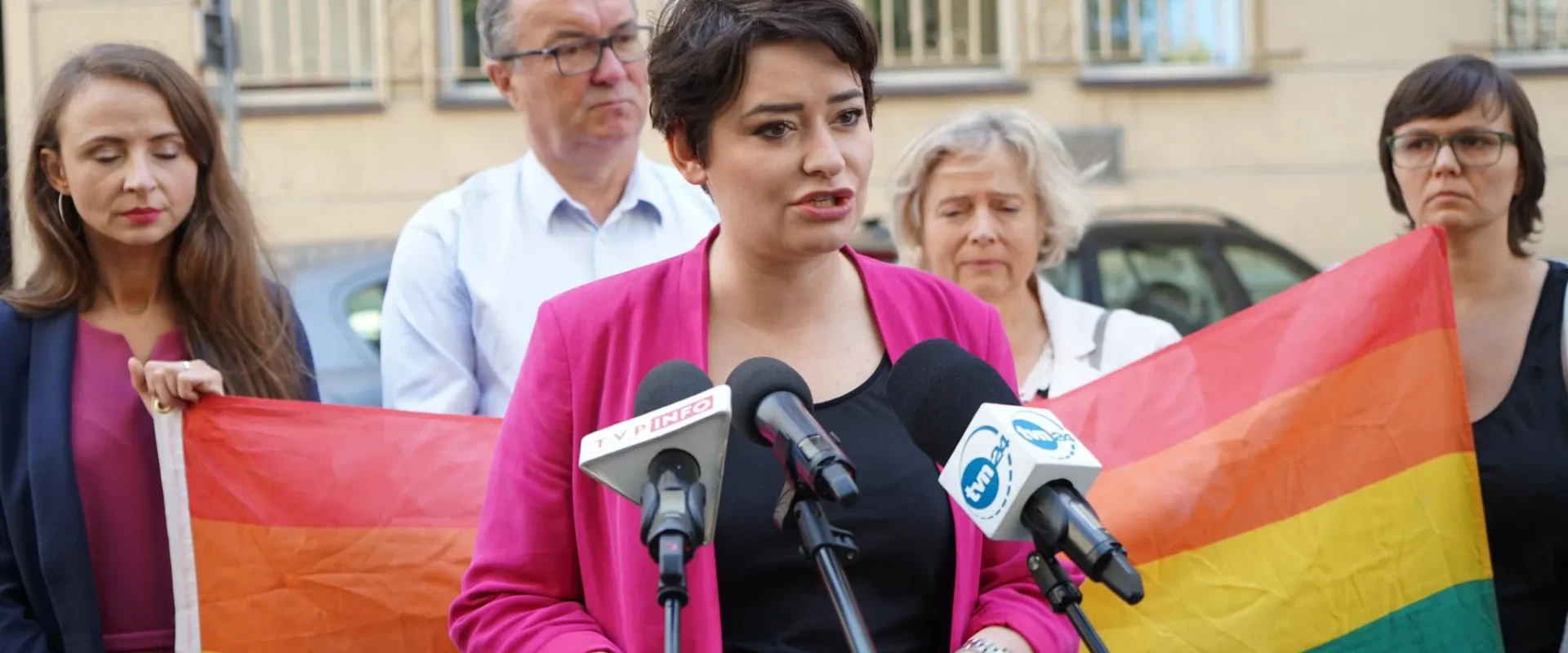 Kobieta w różowym żakiecie przemawia do mikrofonów podczas konferencji prasowej, otoczona innymi osobami, z tęczową flagą w tle.