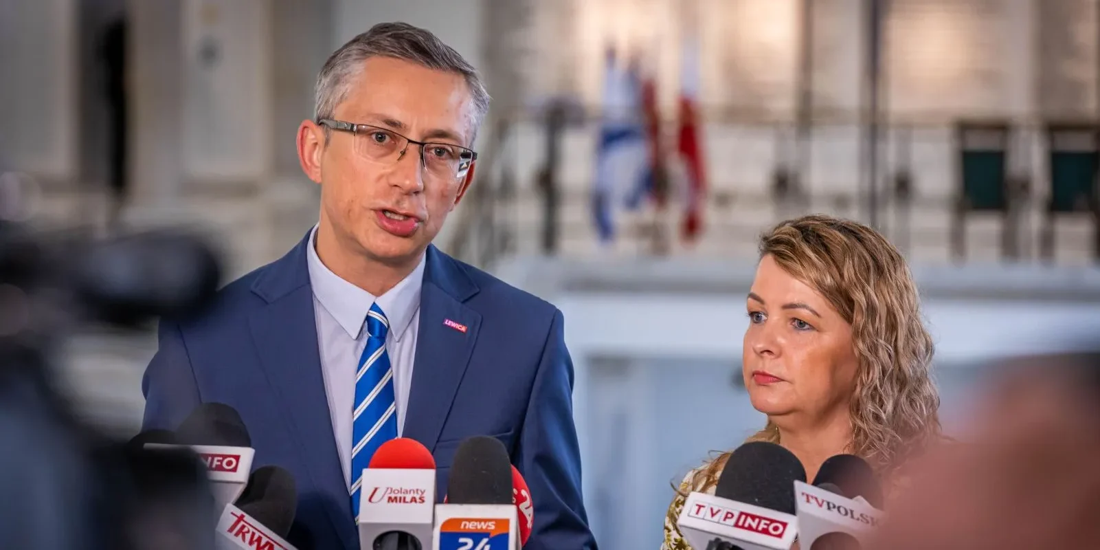Polityk Lewicy w granatowym garniturze i kobieta obok niego udzielają konferencji prasowej przed licznymi mikrofonami i kamerami.