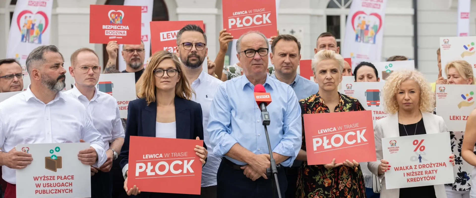 Grupa polityków Lewicy na demonstracji z czerwonymi plakatami, a w centrum mężczyzna przemawia do mikrofonu.