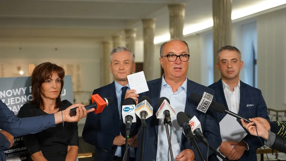 Mężczyzna w garniturze i okularach przemawia do licznych mikrofonów podczas konferencji prasowej, otoczony przez innych polityków i reporterów.