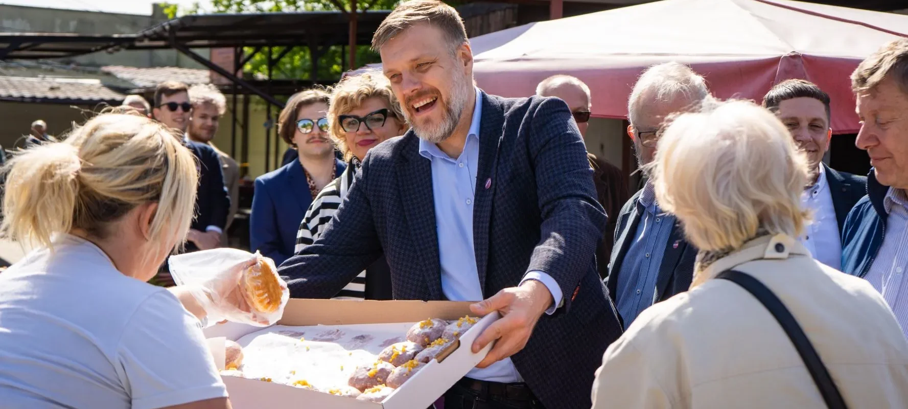 Mężczyzna w granatowej marynarce rozdaje ciastka z pudełka widzom podczas plenerowego wydarzenia w województwie opolskim.