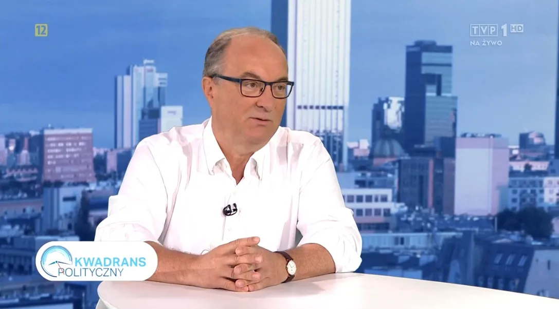 Mężczyzna w białej koszuli i okularach siedzi przy stole w studiu telewizyjnym, z panoramą miasta w tle i logiem programu Kwadrans Polityczny (na żywo).