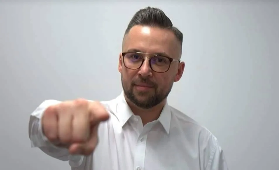 Mężczyzna w białej koszuli i okularach patrzy prosto w kamerę i wyciąga dłoń, wskazując palcem na widza.