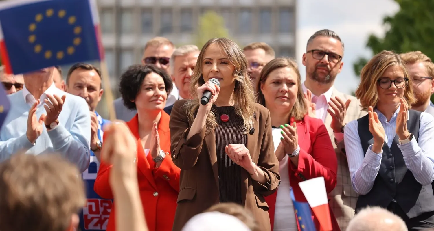 Kobieta przemawia do mikrofonu podczas zgromadzenia, otoczona przez innych uczestników; w tle widoczne flagi Unii Europejskiej i Polski.