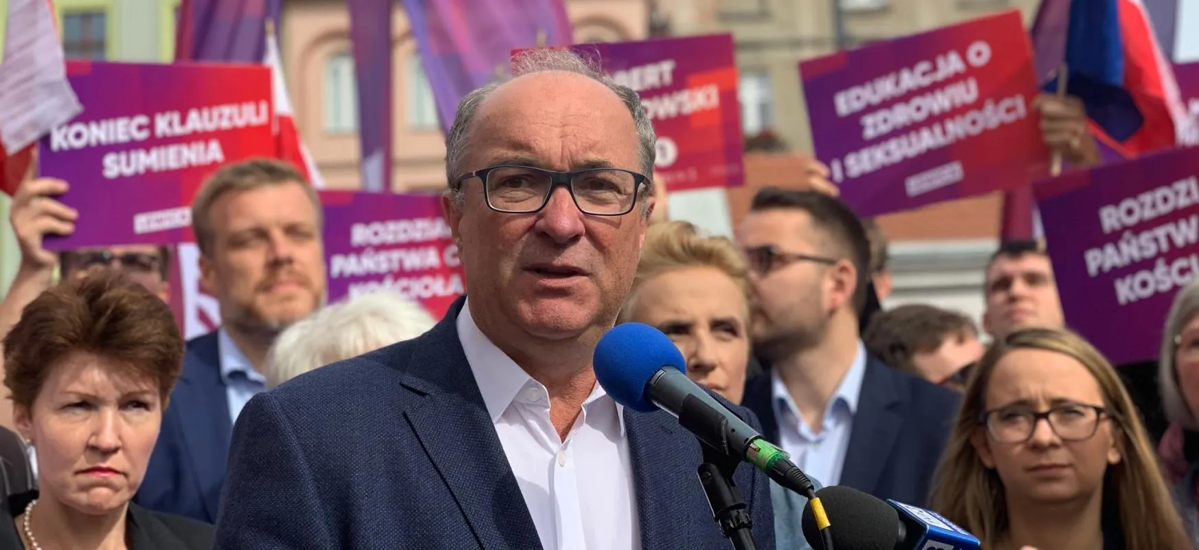 Mężczyzna w garniturze i okularach przemawia do mikrofonu podczas plenerowej konferencji prasowej, wokół niego zgromadzony tłum oraz wielobarwne plakaty w tle.
