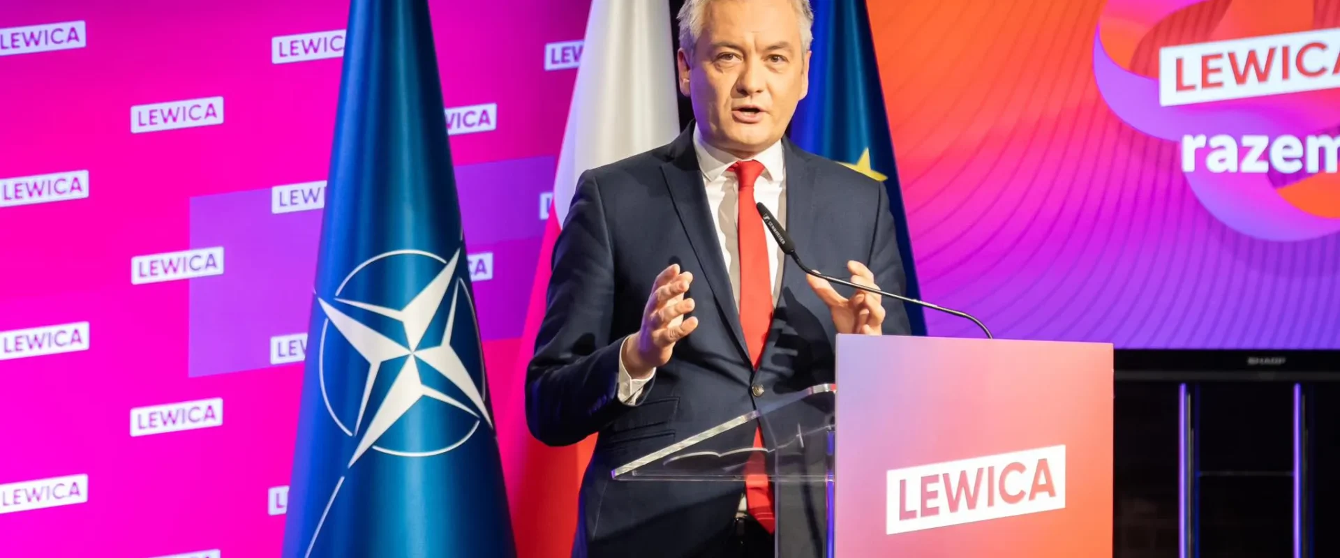 Na zdjęciu widoczny jest mężczyzna przemawiający z mównicy, na tle flag oraz kolorowych bannerów z logo Lewicy.