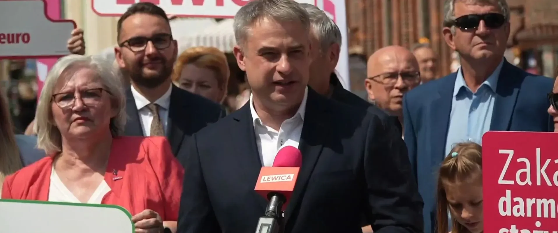 Polityk Lewicy przemawia do mikrofonu podczas plenerowego zgromadzenia, wokół niego ludzie trzymają plakaty i banery z napisem LEWICA.