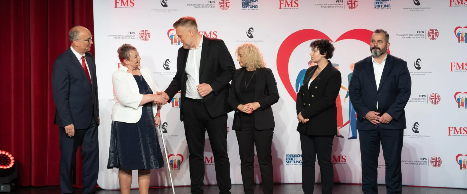 Szóstka osób stoi na scenie podczas wydarzenia; starsza kobieta w białej marynarce ściska dłoń wysokiemu mężczyźnie w czarnym garniturze, a pozostali obserwują, z tyłu biało-czerwony baner z logotypami i dużym czerwonym sercem.