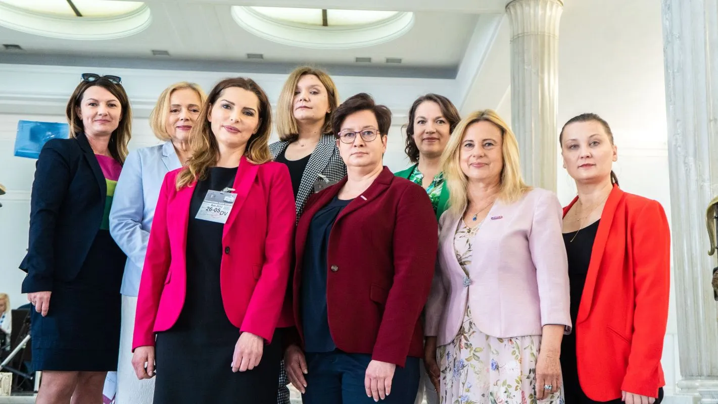 Grupa kobiet w kolorowych marynarkach stoi razem w jasnej sali z kolumnami.