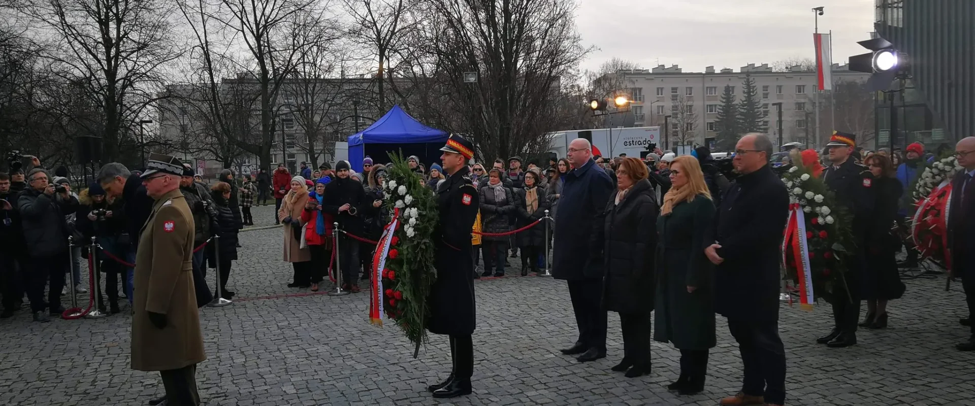Uczestnicy uroczystości upamiętniającej 75. rocznicę wyzwolenia obozu Auschwitz-Birkenau stoją na kamiennym placu, trzymając wieńce i kwiaty, w otoczeniu zgromadzonych ludzi i mundurowych.