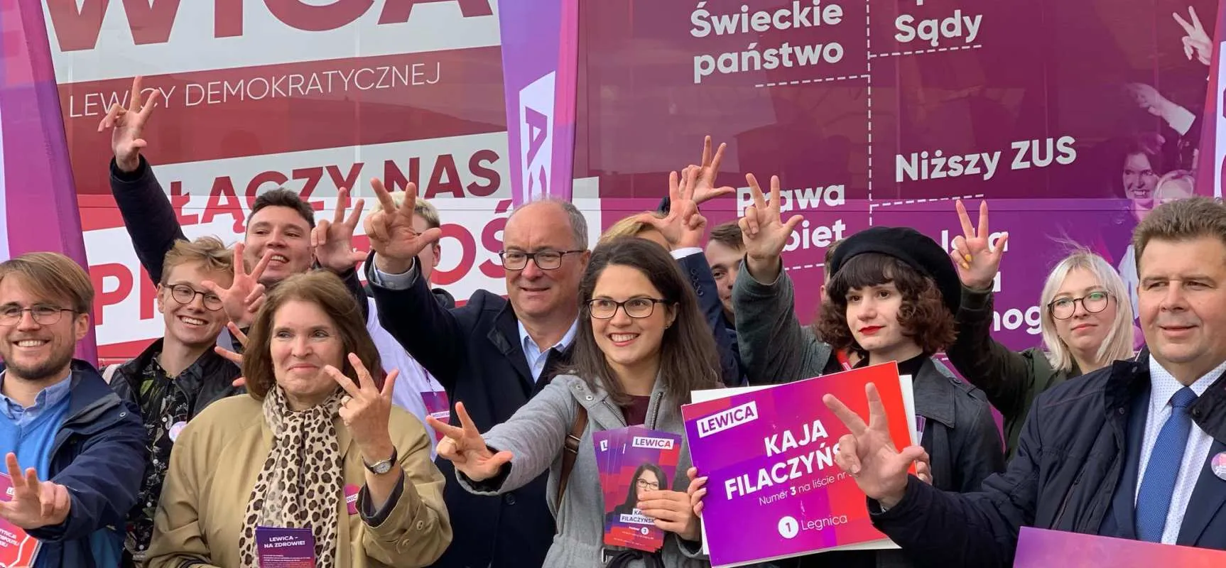 Grupa osób na wiecu Lewicy pozująca do zdjęcia z plakatami i różowymi banerami w tle, pokazująca gest dłoni w kształcie „V”.