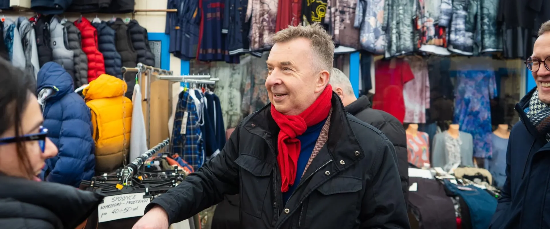 Uśmiechnięty mężczyzna w czarnym płaszczu i czerwonym szaliku rozmawia z innymi klientami na stoisku z odzieżą na targu w Zachodniopomorskiem.
