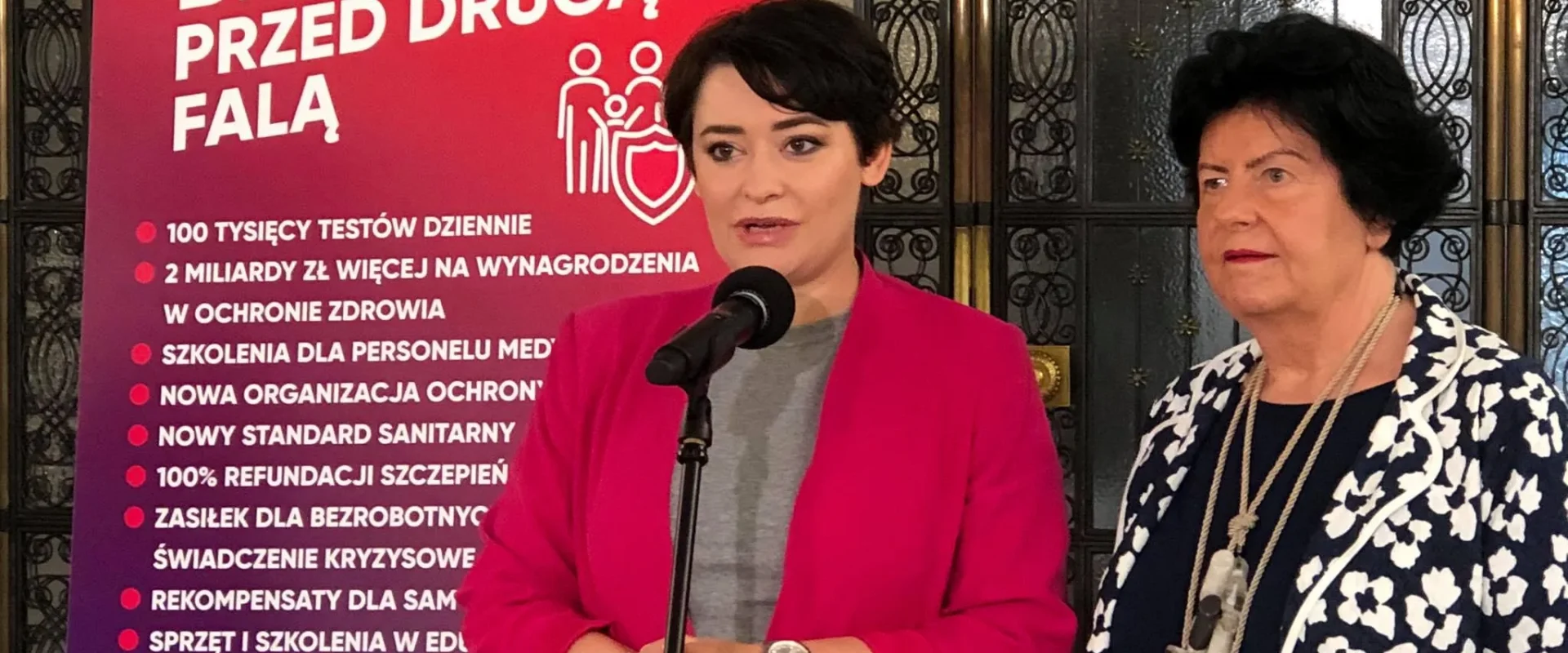 Dwie kobiety stoją przy mikrofonie, na tle dużego plakatu z hasłem „Bezpieczni przed drugą falą” i listą punktów.