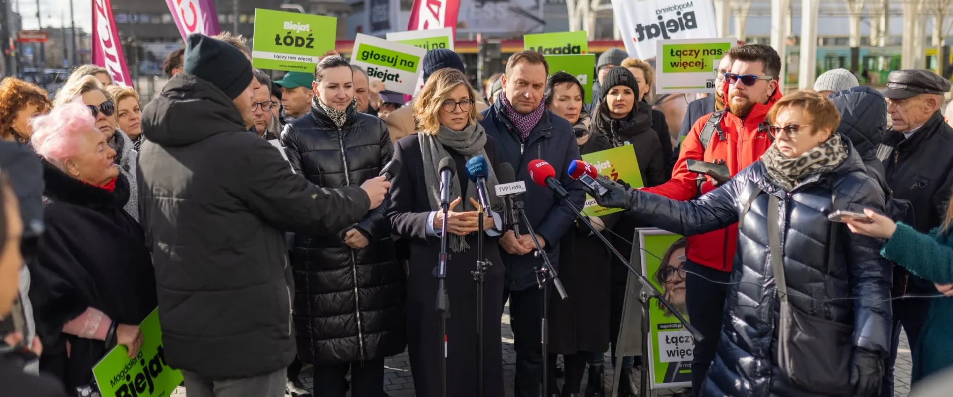 Grupa ludzi na plenerowej konferencji prasowej na placu miasta trzyma zielone plakaty z hasłami popierającymi budowę mieszkań na wynajem i udziela wywiadów mediom.