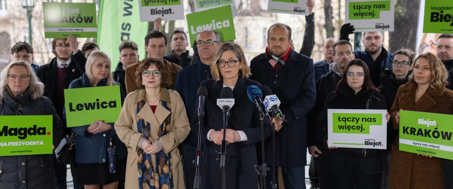 Kobieta w okularach przemawia do mikrofonów podczas konferencji prasowej, otoczona grupą zwolenników trzymających zielone plakaty z napisem Lewica.
