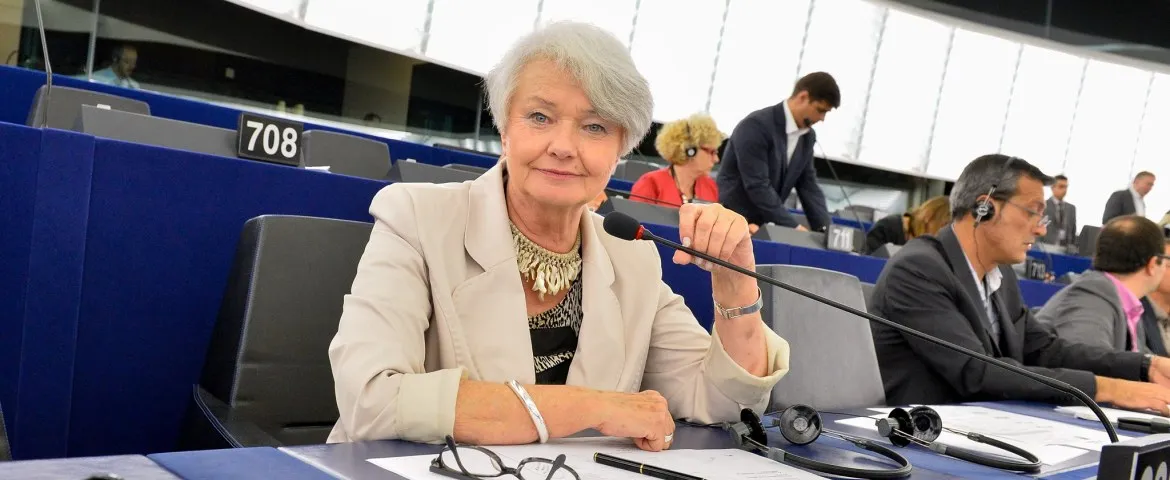 Kobieta o krótkich siwych włosach siedzi przy stole w sali obrad, mówi do mikrofonu, a w tle pracują inni uczestnicy.