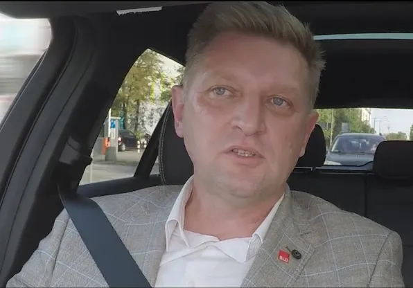 Mężczyzna w jasnym garniturze siedzi w samochodzie, zapiętym pasem bezpieczeństwa, i zwraca się do kamery.