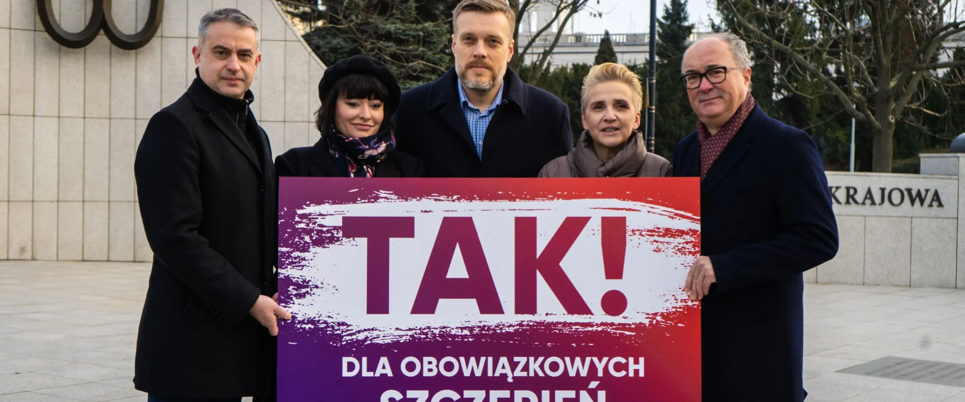 Pięć osób stoi na placu i trzyma duży gradientowy plakat z napisem TAK! DLA OBOWIĄZKOWYCH SZCZEPIEŃ (LEWICA), na tle widoczny pomnik i przestrzeń miejska.