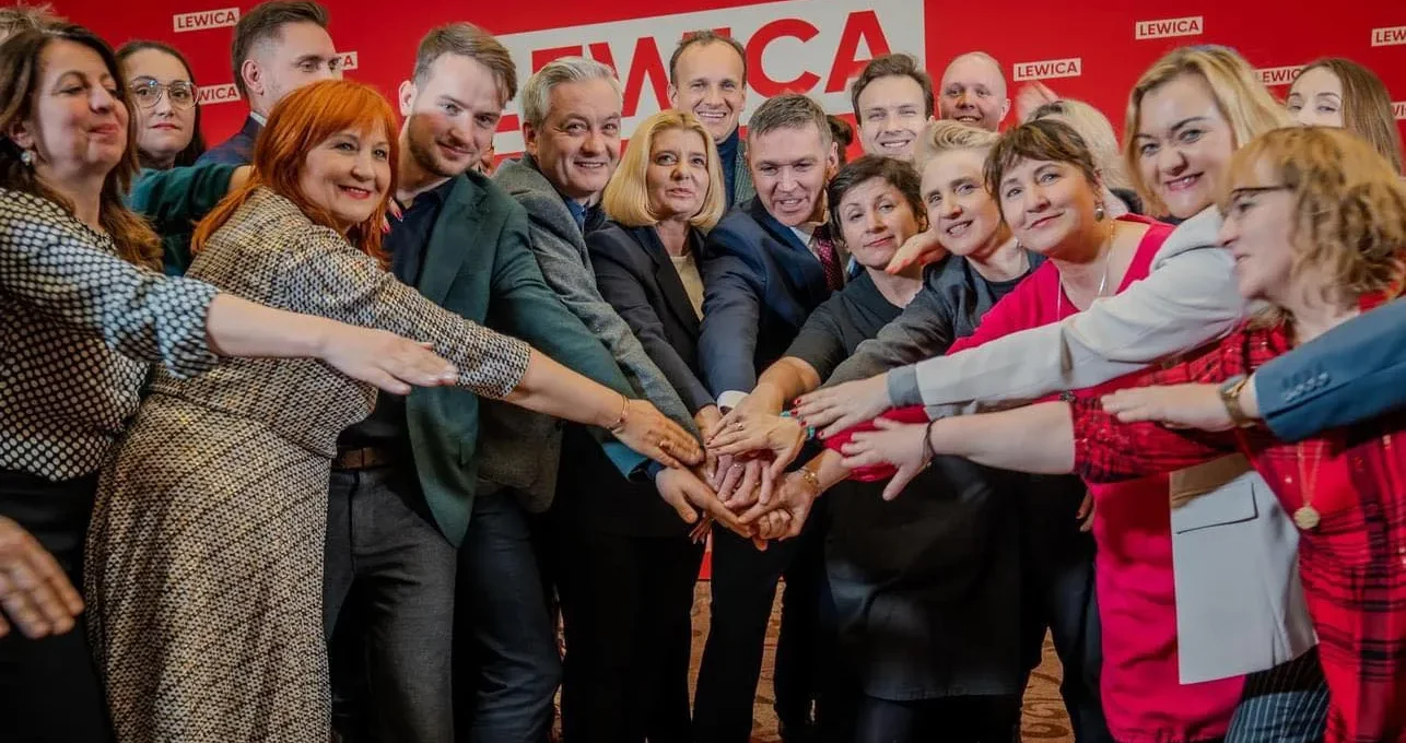 Grupa polityków Lewicy złączają dłonie w geście jedności na czerwonym tle z logiem Lewicy.