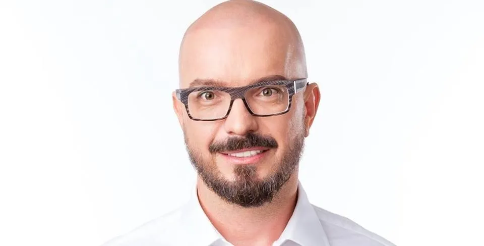 Portret mężczyzny z brodą i okularami, z gładko ogoloną głową, uśmiechnięty, w białej koszuli na białym tle.