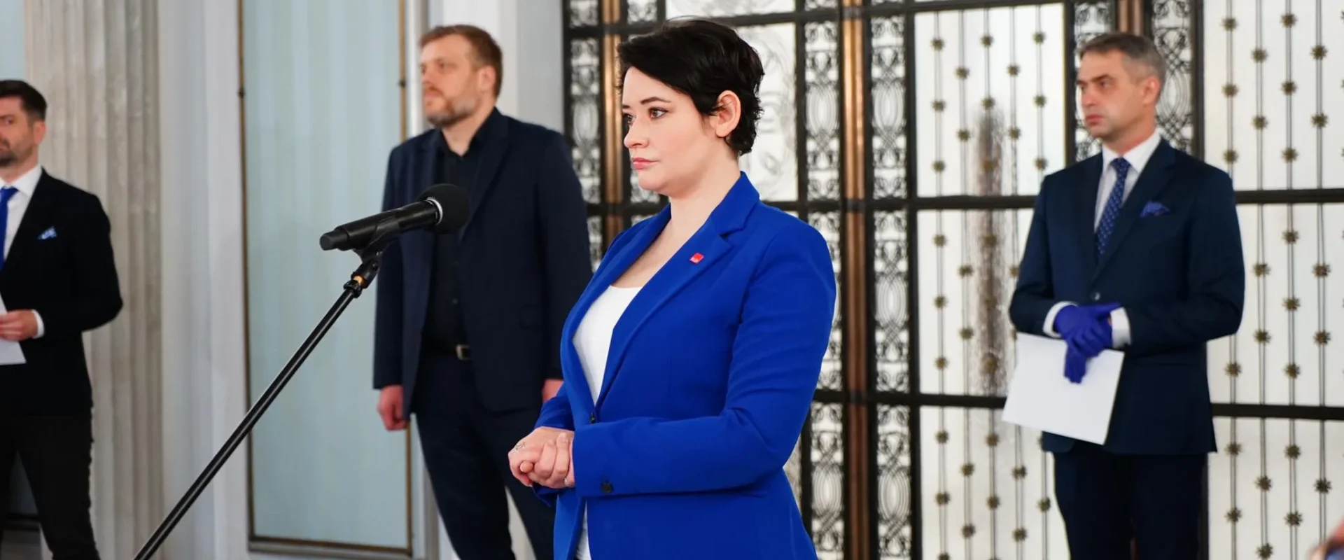 Kobieta w niebieskim żakiecie stoi przy mikrofonie podczas konferencji prasowej, a w tle stoją mężczyźni w garniturach i ozdobne metalowe drzwi.