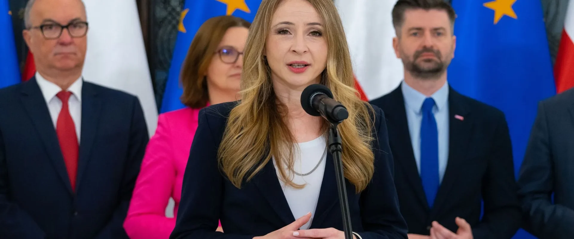 Kobieta w granatowym garniturze przemawia do mikrofonu podczas konferencji prasowej, w tle stoją inni politycy i flagi Unii Europejskiej.